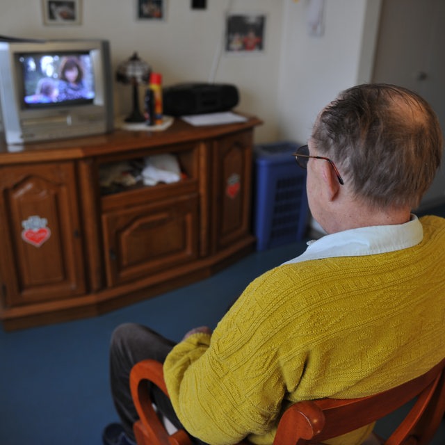 Eine älterer Mann sitzt vor einem Fernseher.