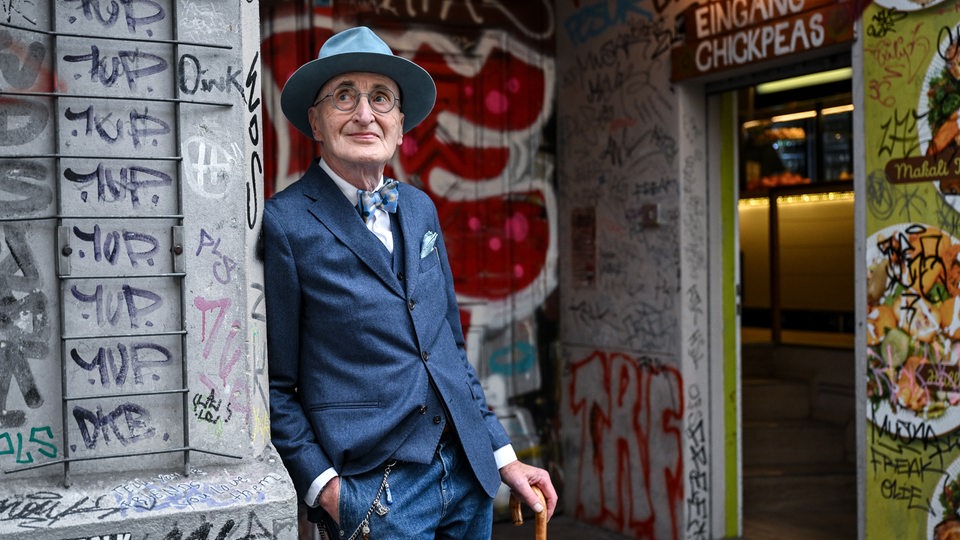 Günther Krabbenhöft mit Stock und Hut,angelehnt sich an eine mit Graffiti bemalte Mauer