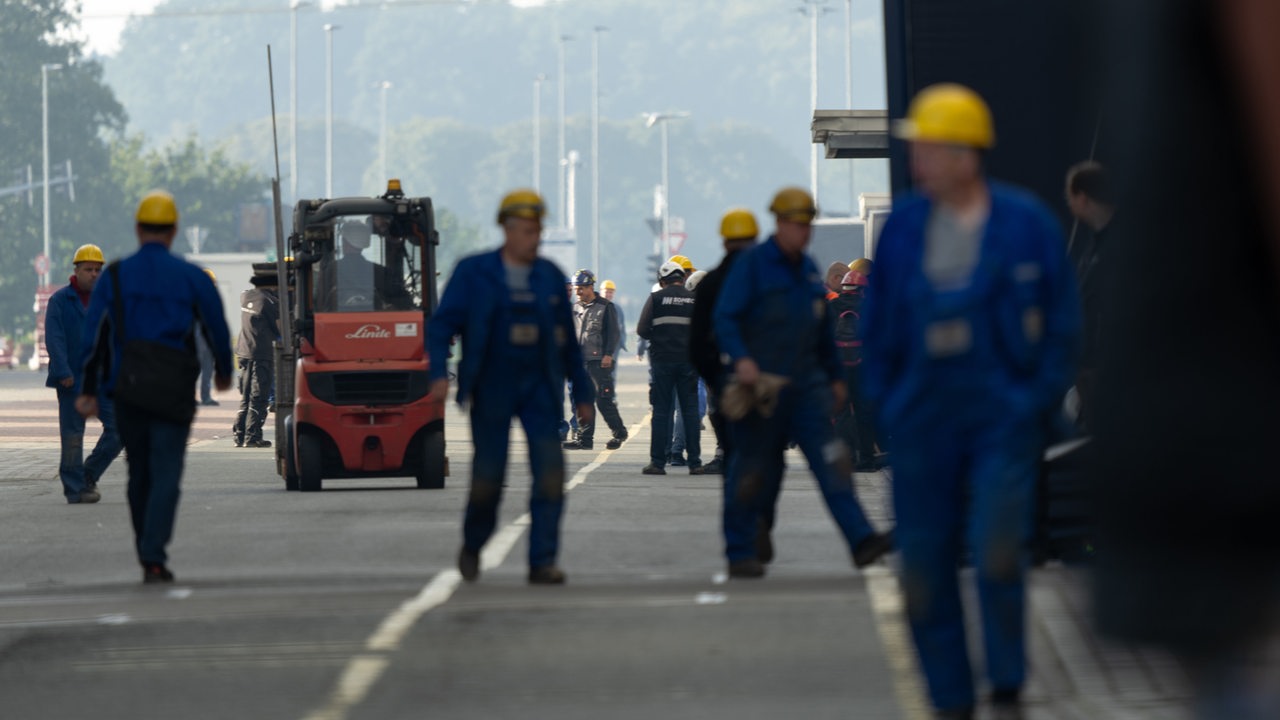 Arbeiter in blauen Overalls und mit gelben Helmen gehen über ein Betriebsgelände.